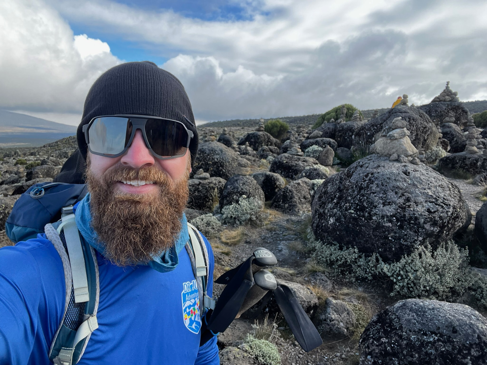 Cole Kline smiling in a beanie and sunglasses on a rocky mountain slope, hiking poles in hand