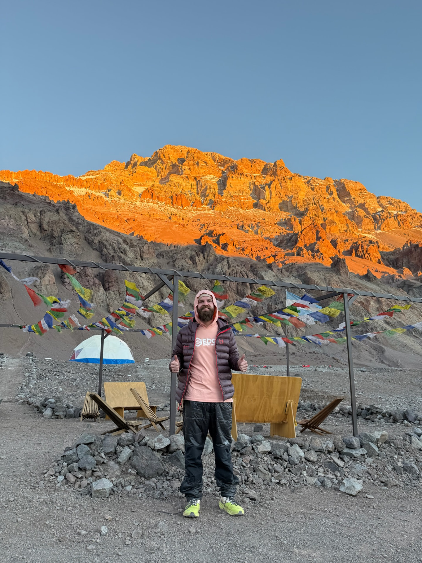 Cole Kline giving two thumbs up at a high mountain camp, prayer flags and a sunlit peak behind him