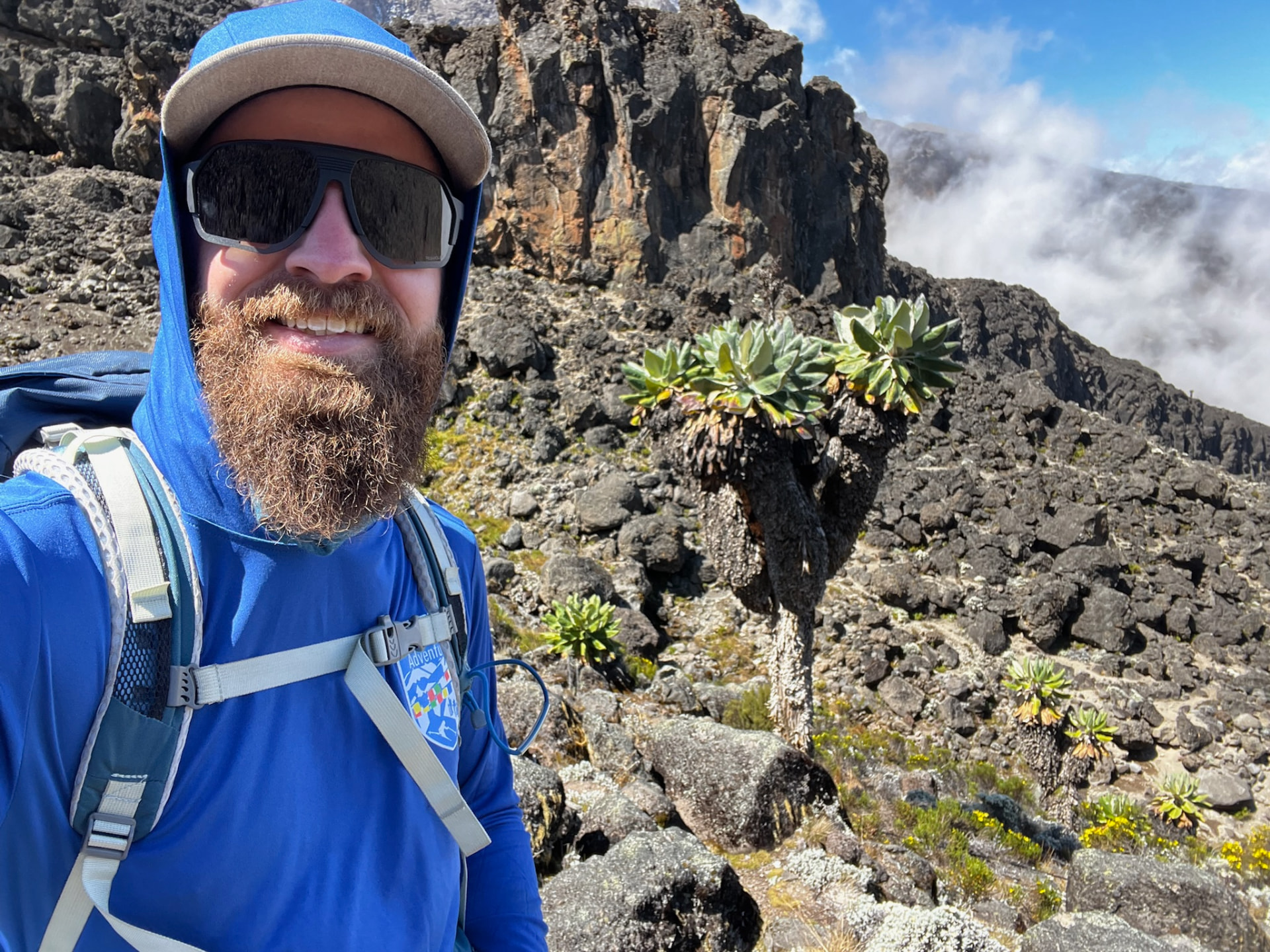 Cole Kline on a rocky mountain trail, smiling in sunglasses and a blue hooded jacket